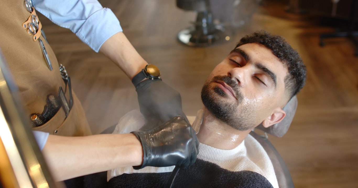 Kunde læner sig tilbage i barberstol med barberkappe, barber udfører barbering med ragekniv til soignering