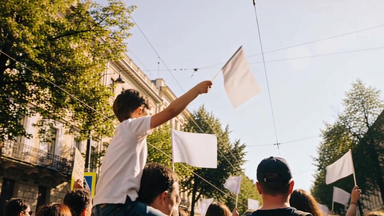Bambino che saluta con una maglietta bianca, seduto sulla spalla di un adulto e che tiene in mano un foglio, mentre inizia una marcia di protesta.