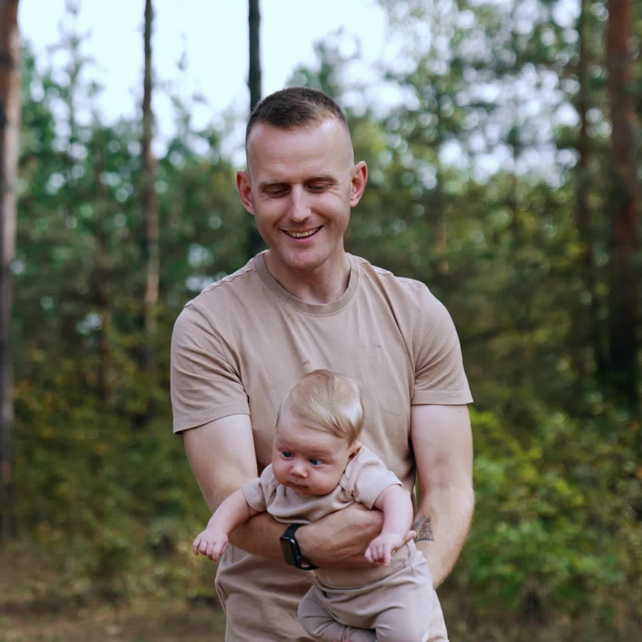 Mann mit beigem T-Shirt winkt mit seinem kleinen Sohn in den Händen. Glücklicher Vater verbringt Zeit mit seinem Sohn in der Natur