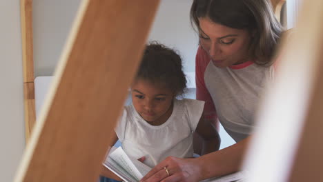 madre y hija hispanas sentadas en la cama de las niñas en casa leyendo un libro juntas