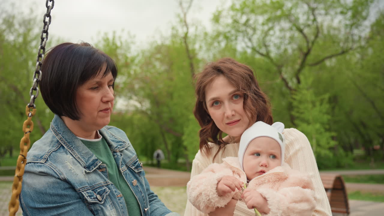 White Mother And White Grandmother Holding Baby On Bench In Leafy Park Smiling Into Camera With Warm Family Embrace Intergenerational Closeness, Denim Jacket, Cosy Baby Outfit And Supportive Presence