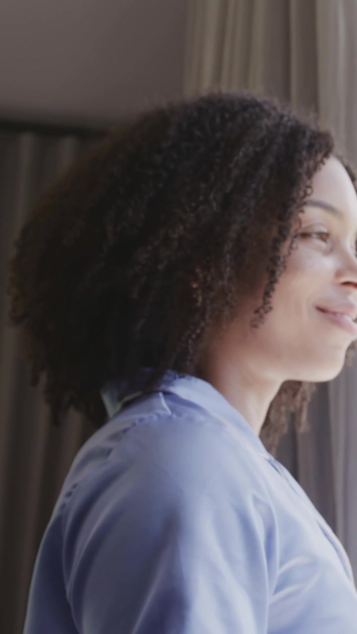 Vertical video: Woman in blue shirt turning into view at window to look out, curtains, copy space