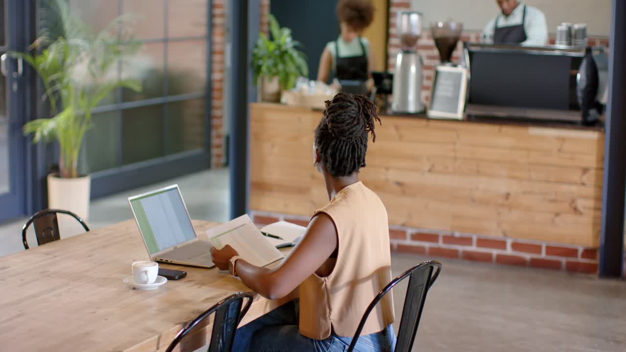 Mulher afro-americana em uma mesa com laptop lendo documentos, observando a equipe se movimentar e acenando para fazer um pedido.