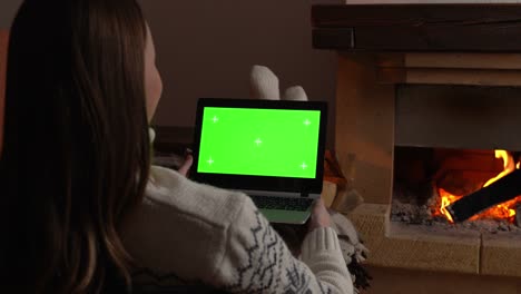 woman lying on sofa in front of fireplace holding laptop green screen, watching chroma key screen by the fireplace