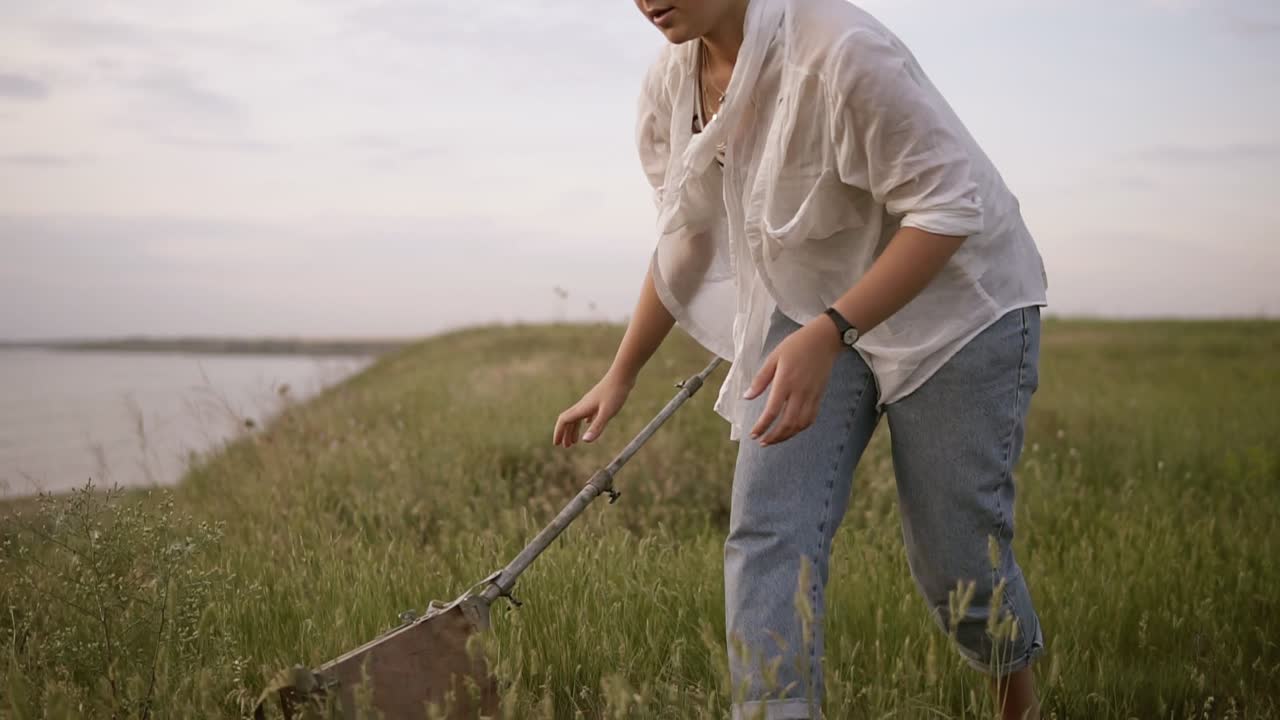 Young brunette girl in white shirt and jeans on meado near the lake. The girl is preparing an easel to work