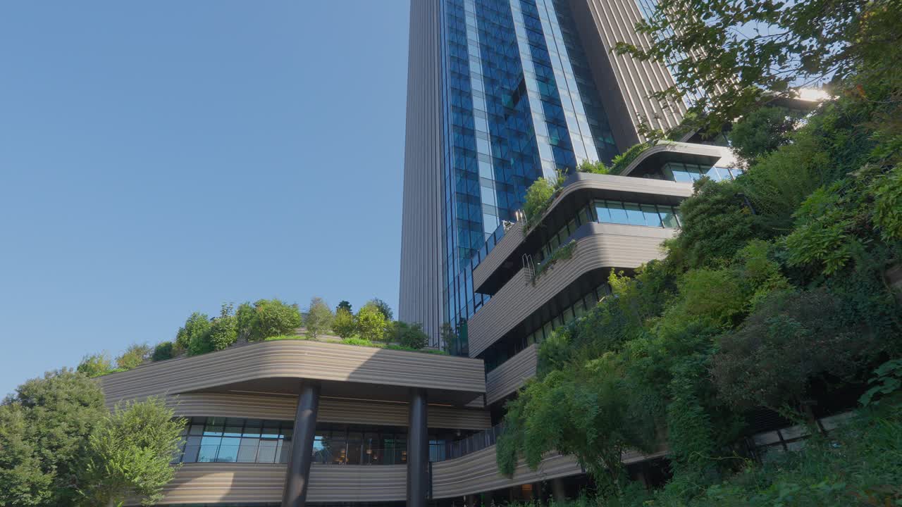 A dramatic low-angle shot of a towering, modern Tokyo skyscraper with striking blue glass and tiered, terraced greenery on its base