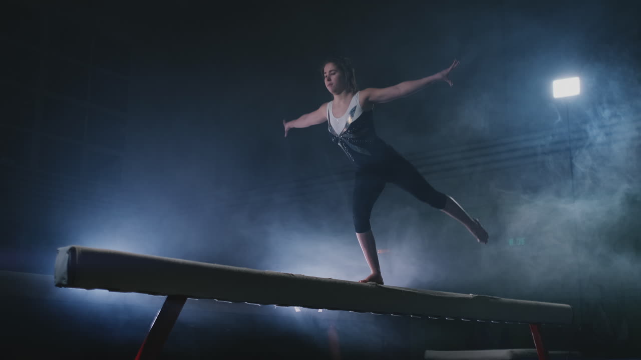 la niña es una atleta profesional realiza un truco acrobático de gimnasia en una viga con luz de fondo y cámara lenta en ropa de gimnasia deportiva. humo y azul. saltar y girar en la viga de equilibrio