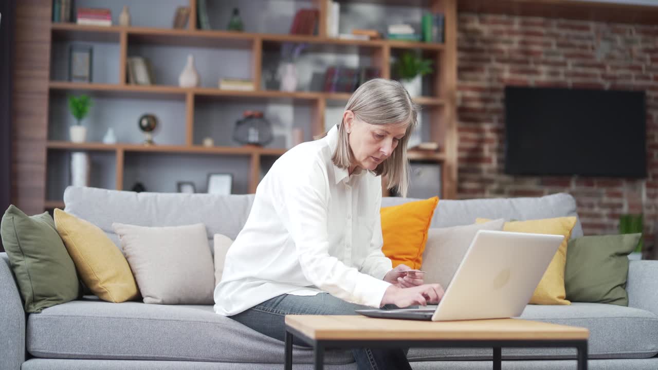 una mujer anciana de cabello gris paga en línea con tarjeta de crédito usando una computadora portátil, ingresa datos, pago de facturas, reserva de boletos, compras.