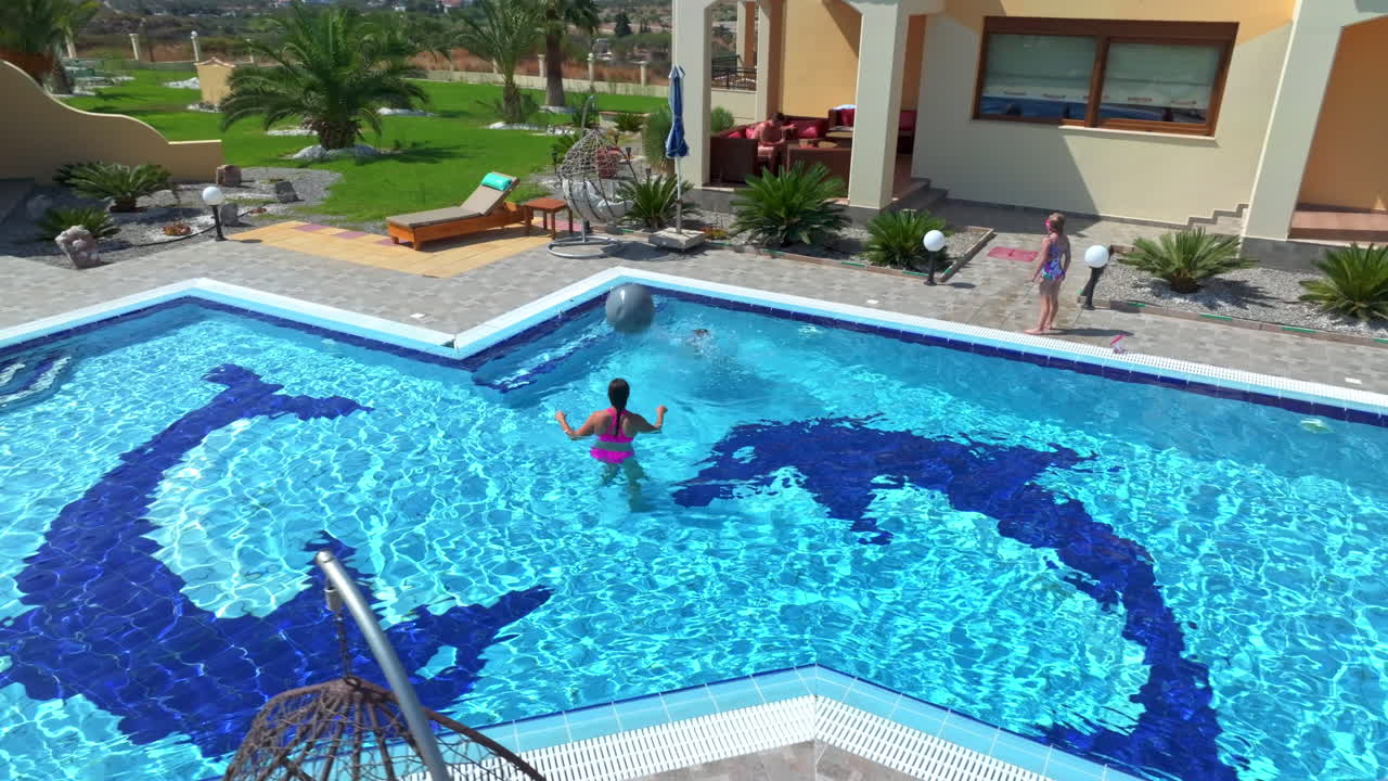 Aerial view orbiting a mom and children playing ball in a villa pool, sunny day