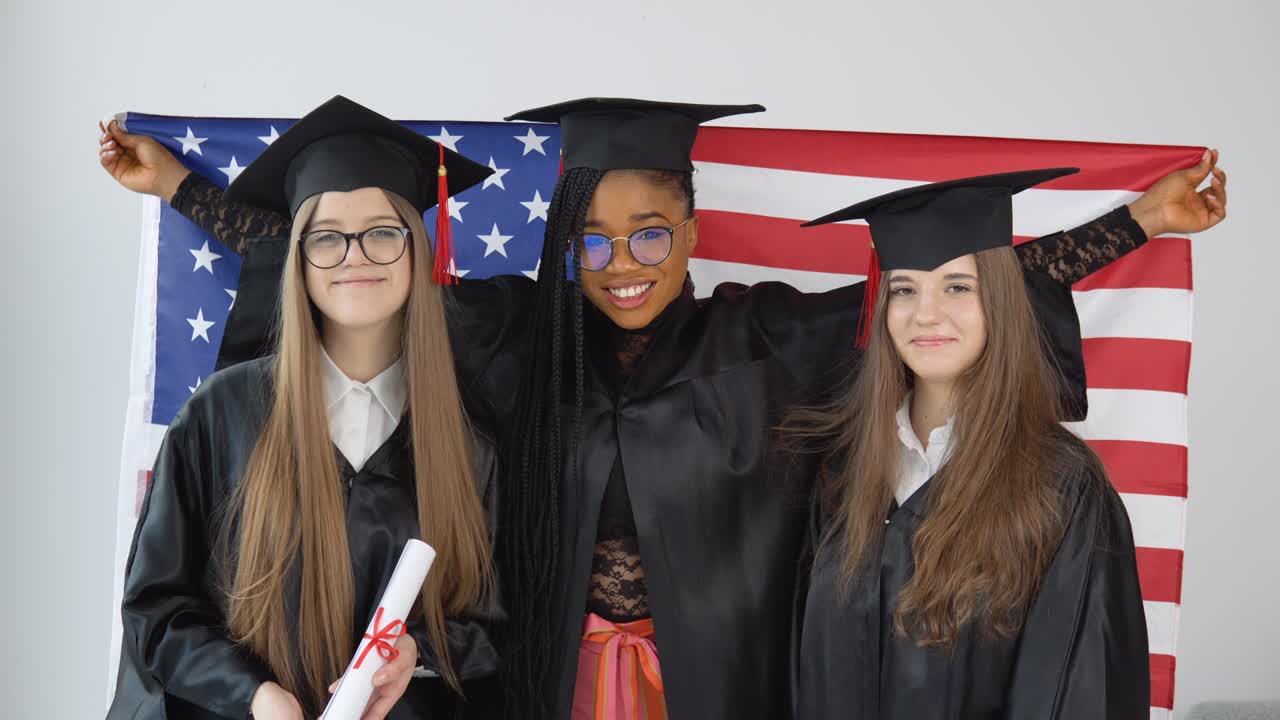tres estudiantes de mujeres en trajes y sombreros de graduación sostienen la bandera de los estados unidos detrás de sus hombros. estudiantes de diferentes razas con la bandera de estados unidos