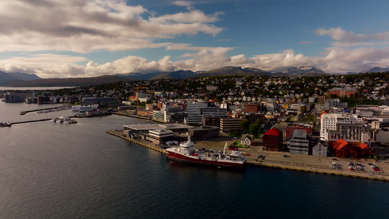 Aerial drone view of Tromso city and harbor in early autumn. Ships dock at the port with colorful buildings and mountains in the background under a cloudy sky in Northern Norway