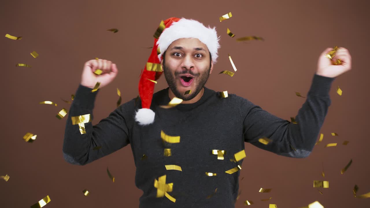 feliz y alegre hombre indio con sombrero de santa lanzando brillo dorado y gritando, disfrutando de las vacaciones de navidad, fondo marrón