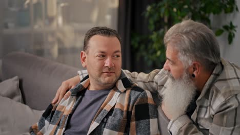 Happy-brunette-man-in-a-checkered-shirt-communicates-with-his-middle-aged-boyfriend-with-gray-hair-and-a-lush-beard-in-a-modern-apartment-on-the-sofa-in-the-evening