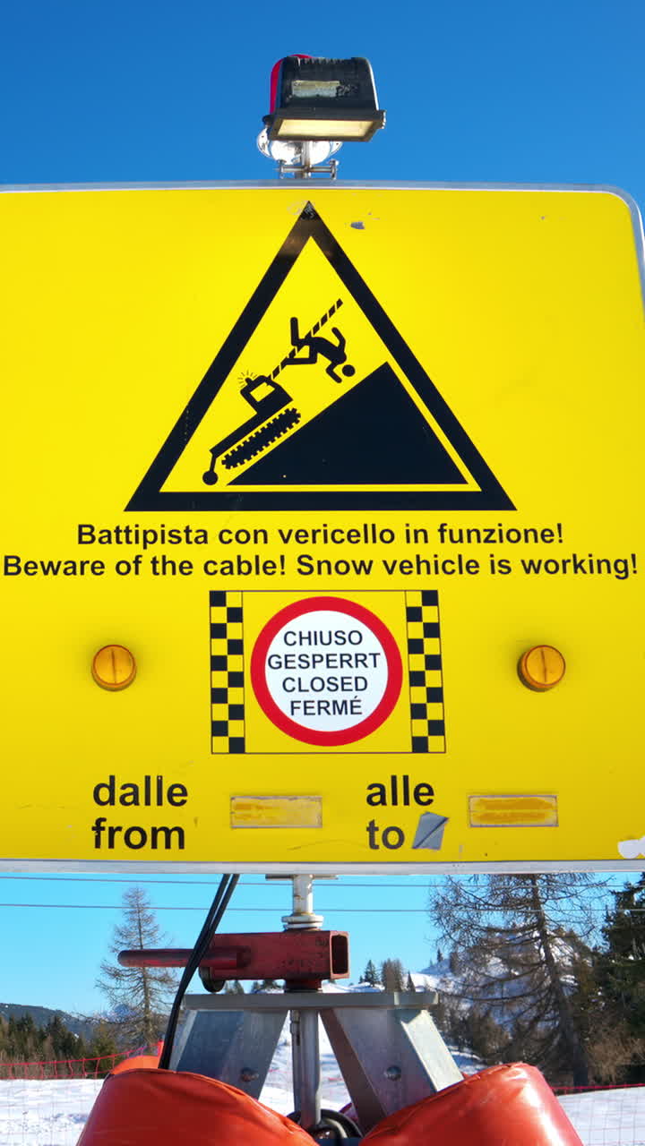 Yellow warning sign about the presence of a potentially dangerous winch cable stretched across the slope on a ski resort. Vertical