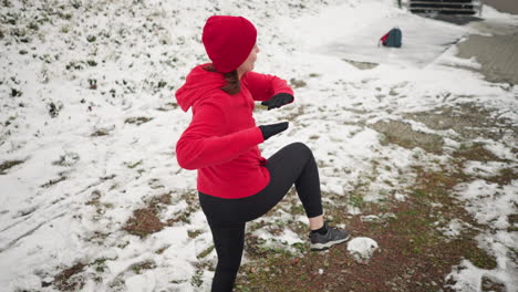 lady performing energetic winter workout outdoors, lifting leg with hand for balance on snowy ground near staircase, surrounded by snow, background features bag and snowy pathway