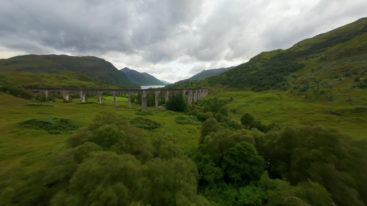 Flyfoto over Glenfinnan Viadukt, svinger til høyre for å vise frem de ikoniske steinbuene med det vidstrakte, dypblå vannet i Loch Shiel som en majestetisk bakgrunn i det skotske høylandet.