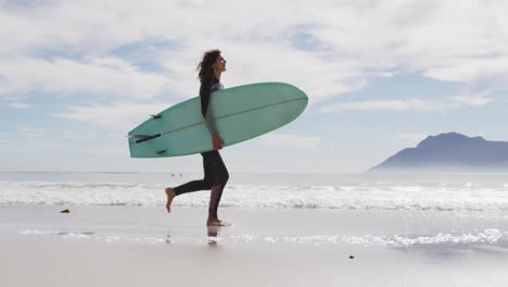 happy mixed race woman running along beach by the sea carrying surfboard