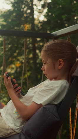 thoughtful child uses contemporary mobile phone sitting in comfy chair. focused girl plays on snug terrace overlooking tranquil lake and untouched woodland