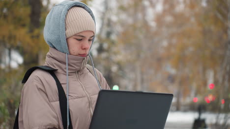 young woman wearing layered winter clothing holds laptop while working outdoors on snow-covered path, focused on screen with blurred background of autumn trees and people walking in distance