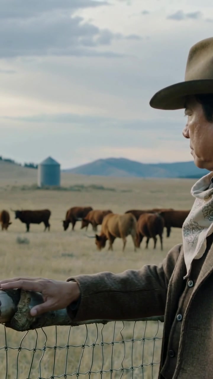 Video verticale: Panoramica di un cowboy con cappello di feltro che fa scorrere la mano lungo una ringhiera mentre controlla la mandria in un ranch, con spazio per il testo.