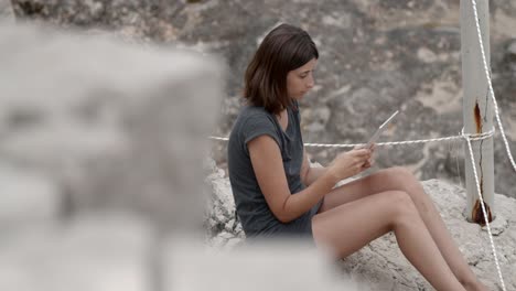 Mädchen-Sitzt-An-Einem-Schönen-Felsigen-Strand-Mit-Tablet-Computer-In-Der-Hand