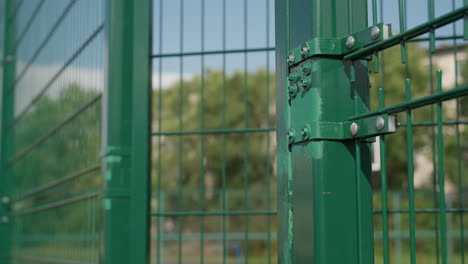 close-up of coach opening green metal gate, volleyball under arm, student following, with lush trees in background, symbolizing entry to sports training