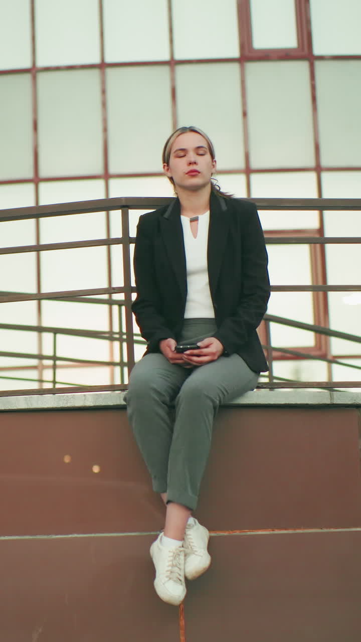 Young girl in black blazer seated on fence in urban area, holding phone while waiting for someone, legs crossed at ankle, modern glass building in background