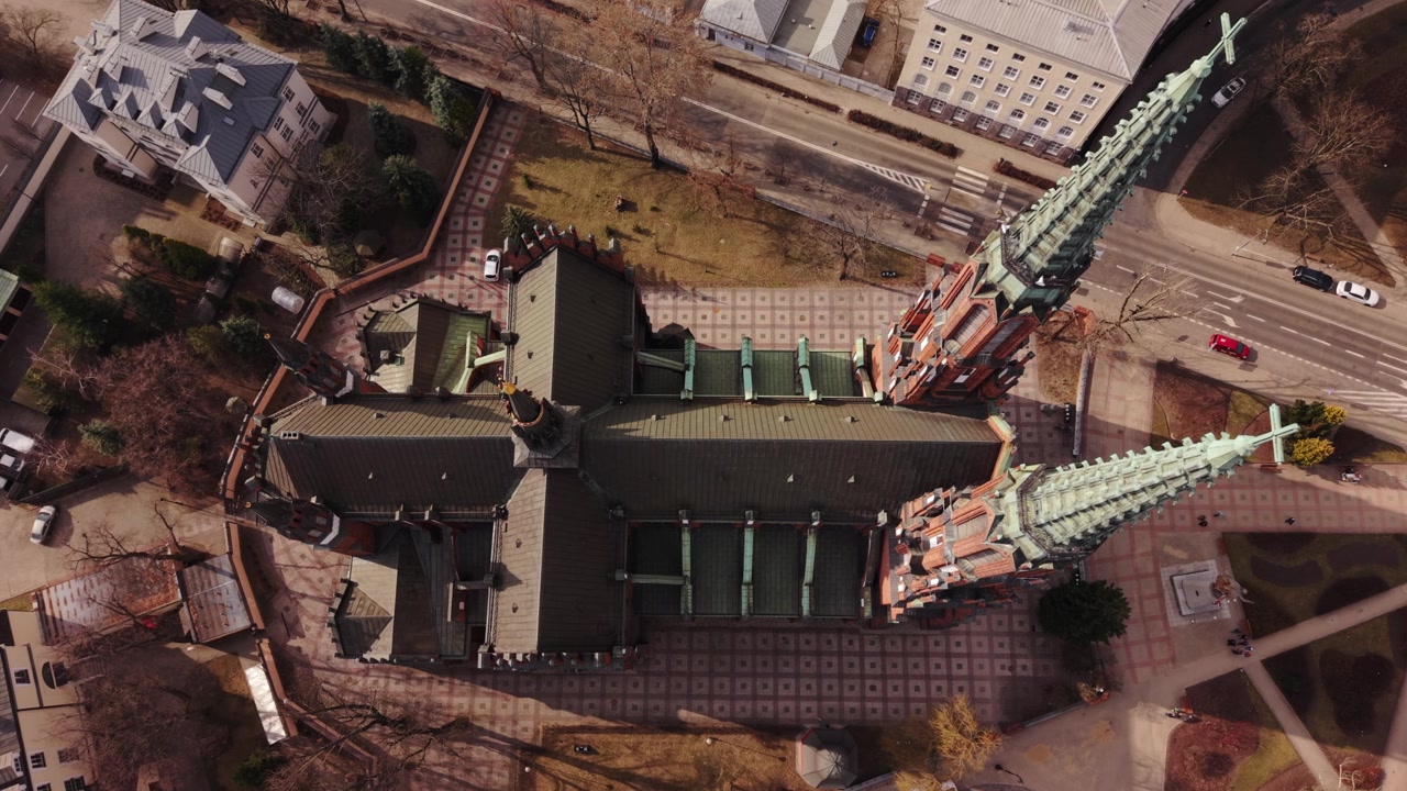 Luftaufnahme einer historischen Kirche in Warschau, Polen, mit Zwillingstürmen, Ziegeldach, Kopfsteinpflasterplatz, geparkten Autos und einer ruhigen Straße. Die Drohnenperspektive von oben betont gotische Details, Kupfer.