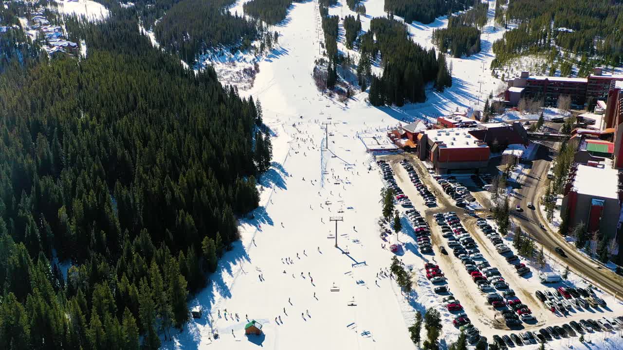 Bird's Eye View of a Ski Trail with People Riding Chairlifts down Slope next to Parking Lot and Pine Tree Forest in Snow