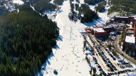 vista de pájaro de una pista de esquí con gente montando telesillas cuesta abajo junto al estacionamiento y un bosque de pinos en la nieve