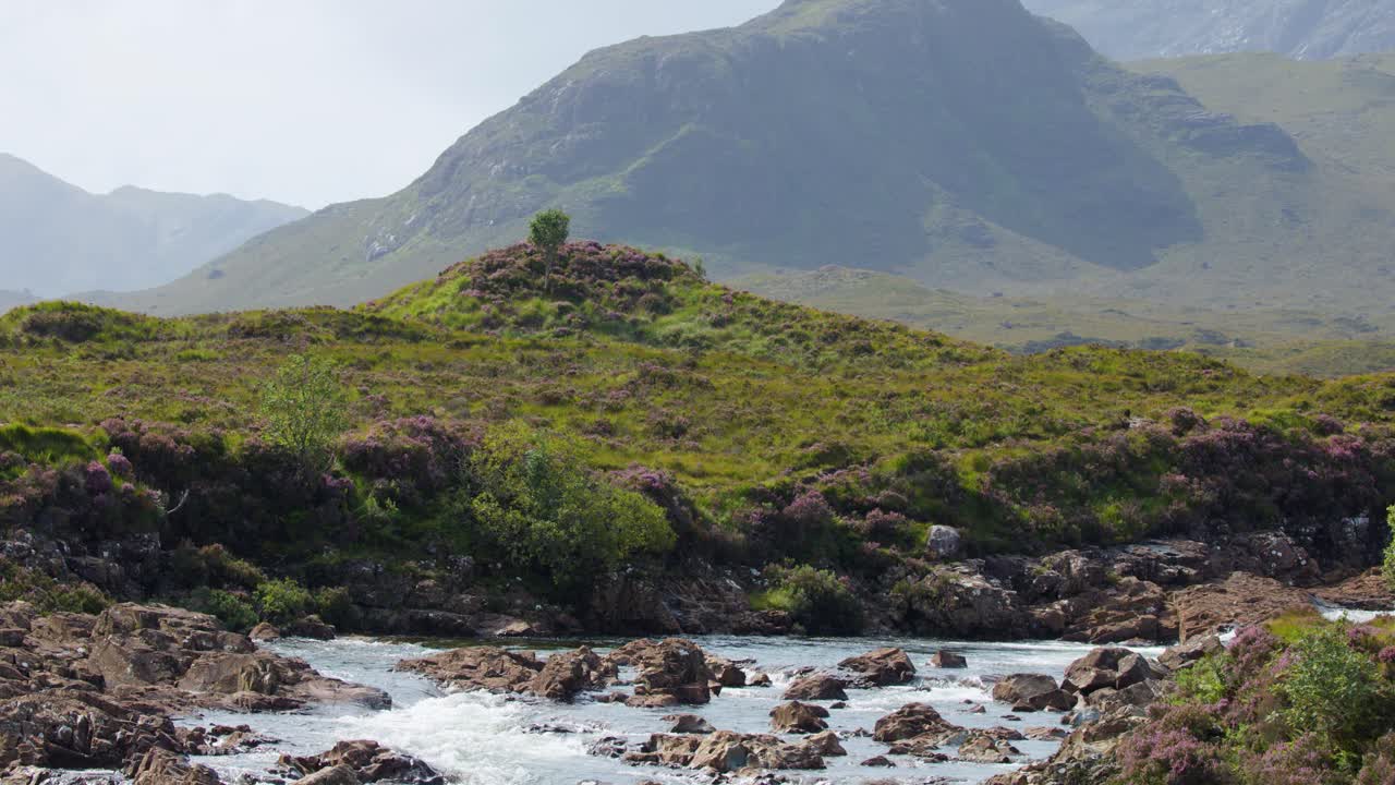 Clear river flows over rocks through green hills, wildflowers, and distant mountains under daylight