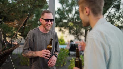 hombre feliz con gafas de sol y barba chasquea botellas marrones con su amigo mientras su novia se acerca y se une a la conversación en el patio trasero mientras se relaja