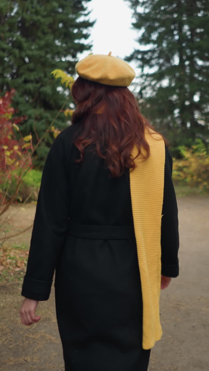 Vista posteriore di una giovane donna con lunghi capelli rossi ondulati, che indossa un cappotto nero, un berretto giallo e una sciarpa abbinata, mentre cammina lungo un sentiero sterrato attraverso un lussureggiante parco verde con un colorato fogliame autunnale.