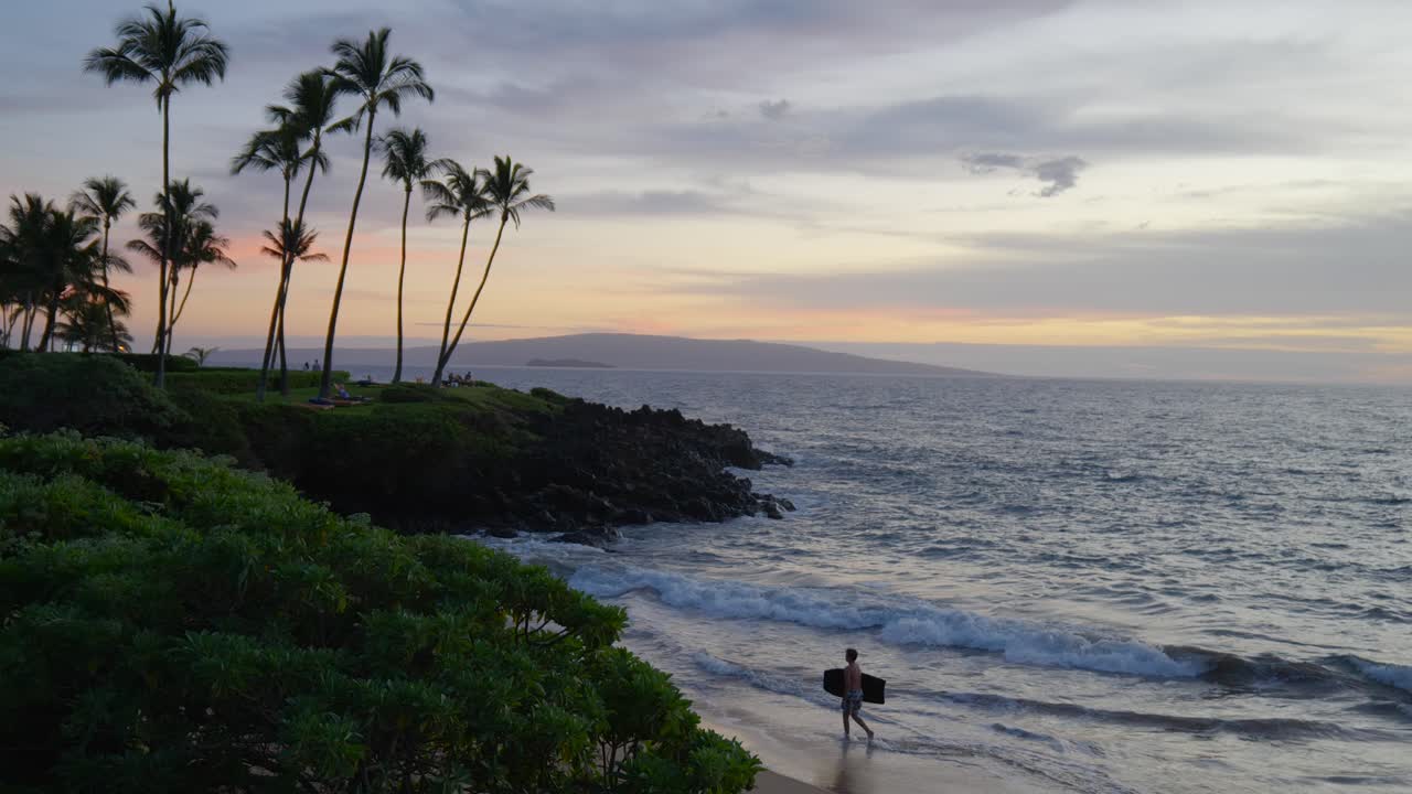 Teenage boy walking along the beach with a body board, at sunset, in Maui Hawaii