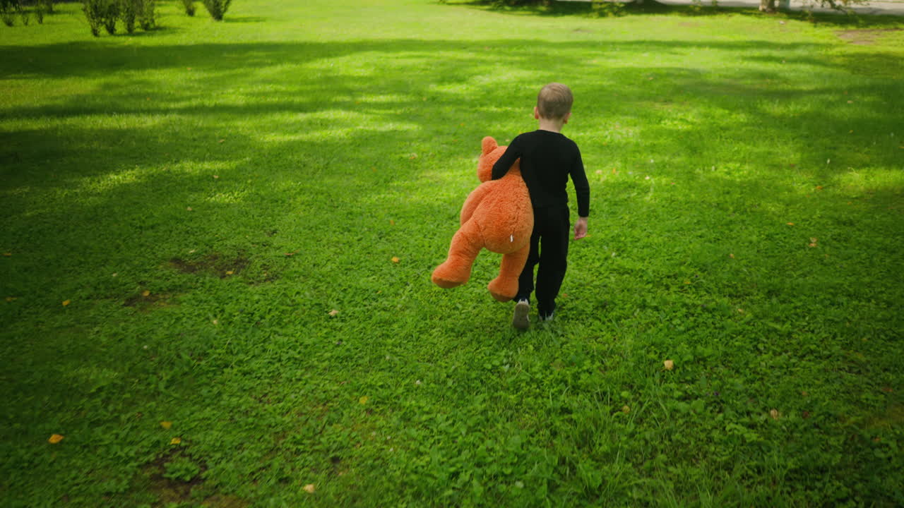 Back view of young boy in black outfit carrying large orange teddy bear while walking across bright green grassy field, surrounded by scattered trees and sunlight patches in peaceful outdoor environment
