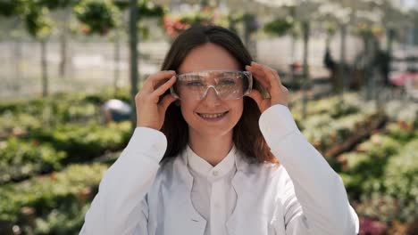 retrato de una mujer joven en una bata blanca con gafas sobre un fondo de un invernadero con plantas