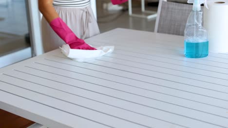 person wearing gloves cleans a table using a spray bottle and cloth in a bright room