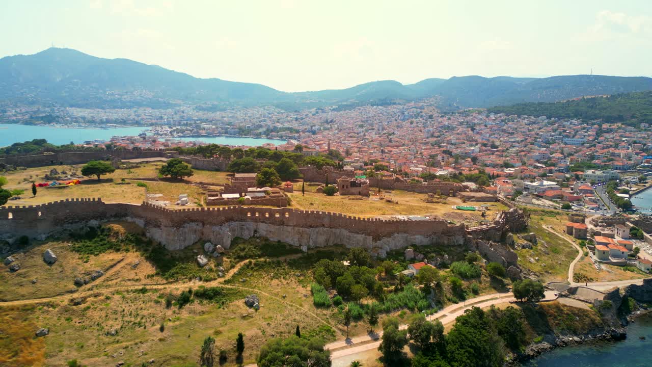 Drone shot of the Castle of Mytilene, Greece,revealing a view of the port, terracotta roofed houses, and mountain hills.