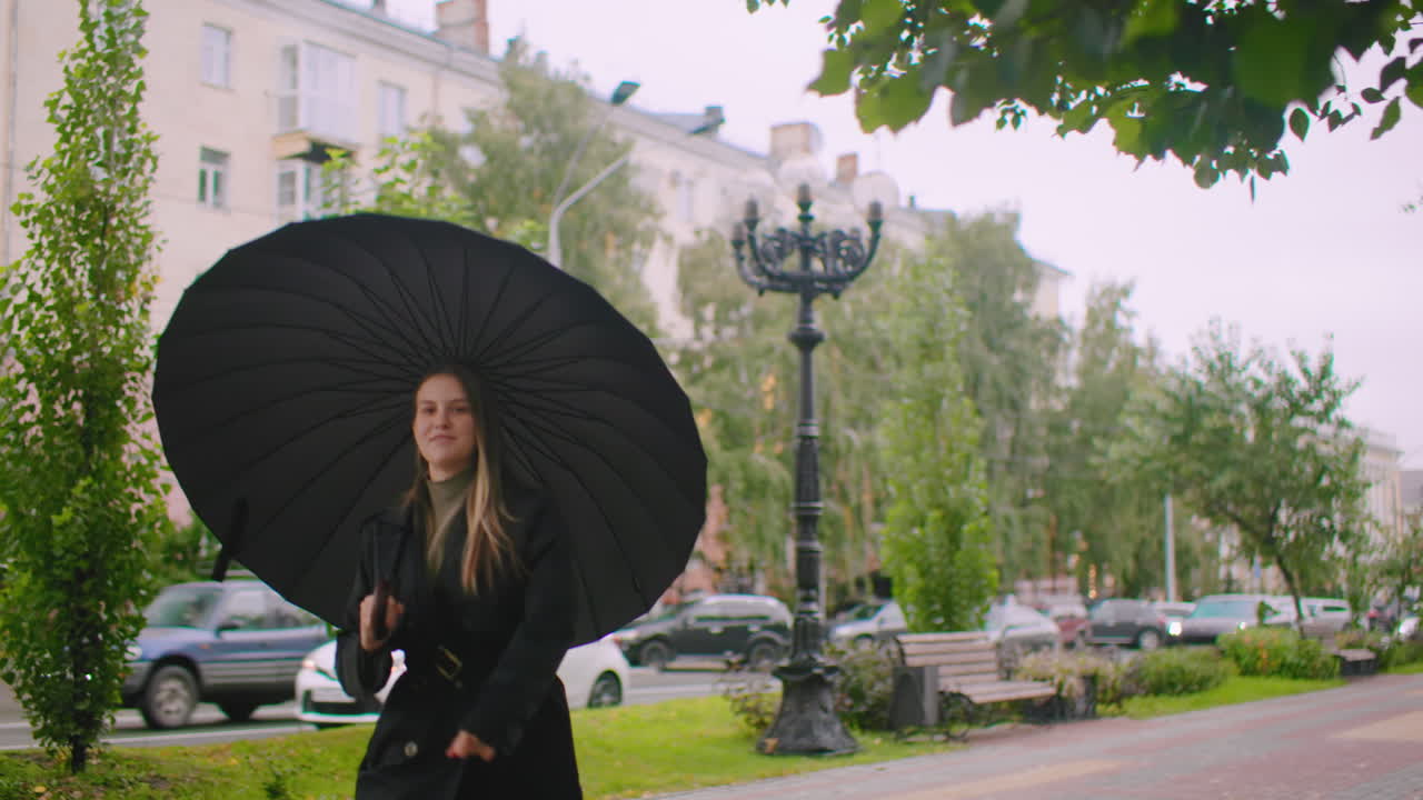 Smiling young woman holding large black umbrella stands on city sidewalk during cloudy day, dressed in black coat, with cars, benches, street lamps, and greenery creating atmospheric urban park scene