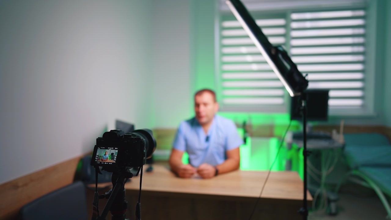 Telecamera da studio che riprende un uomo alla scrivania sotto un'illuminazione ambientale verde. Un rig in primo piano con anteprima posteriore e un presentatore sfocato seduto dietro una scrivania di legno vicino alle tende.