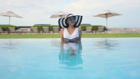 Beautiful-Asian-woman-with-sunglasses-and-striped-hat-sitting-in-a-swimming-pool