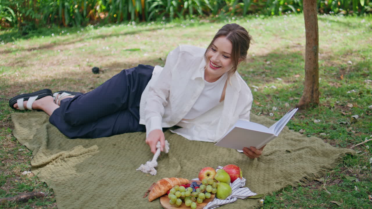 Pretty brunette playing pet lying on picnic blanket with book. Lovely poodle