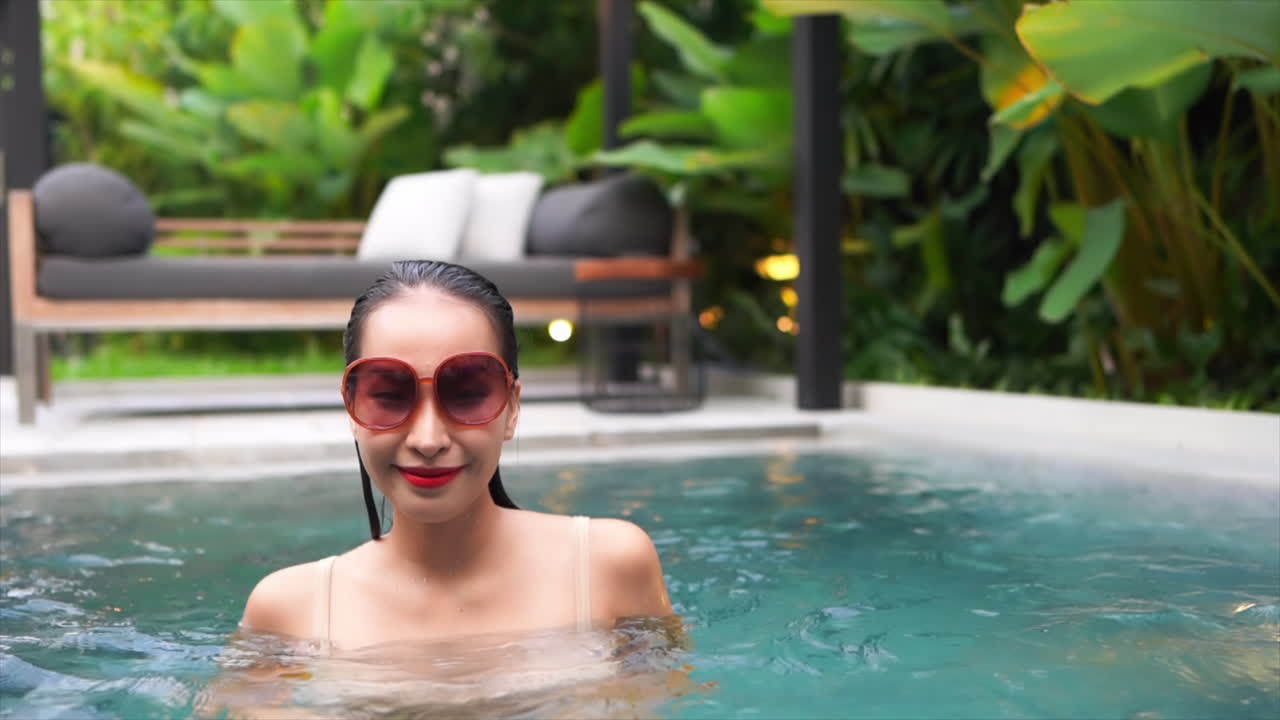beautiful asian girl wearing large sunglasses moving seductively in an inviting swimming pool with crystal clean rippling water with a pool lounge and lots of deep green foliage in the background.