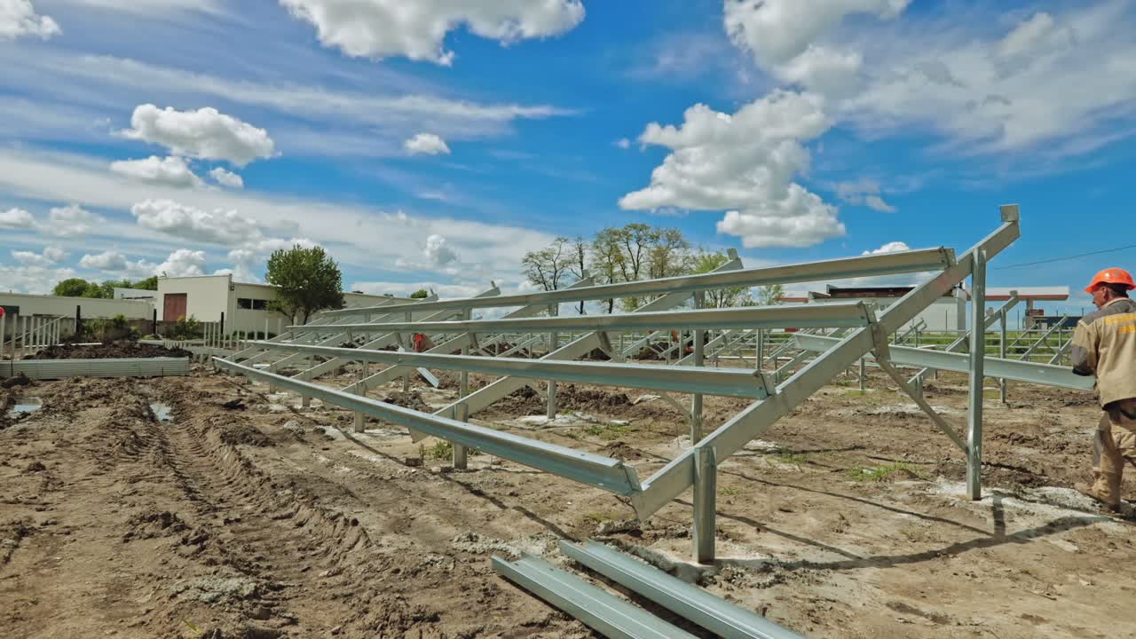 Construction of solar farm on the ground. Workers in uniform and orange helmets build metal basis for solar panels. Time lapse.