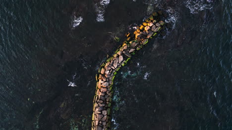 Drone-top-down-rotate-above-moss-covered-wave-break-pile-of-rocks-in-ocean