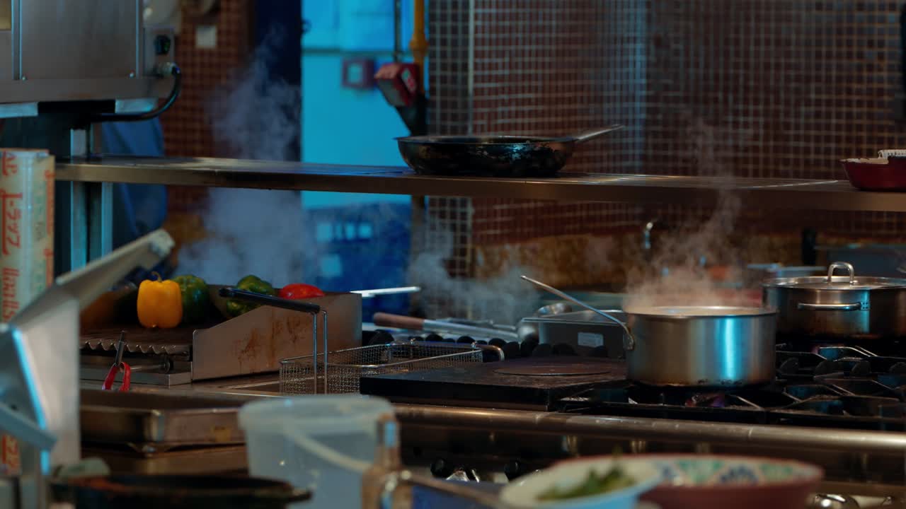 Busy commercial kitchen filled with steam from pots and pans on the stove, fresh bell peppers and other vegetables prepared for cooking