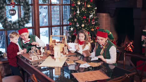 girl and boy are playing at the table with elves in green costumes. santa claus house with christmas tree and fireplace