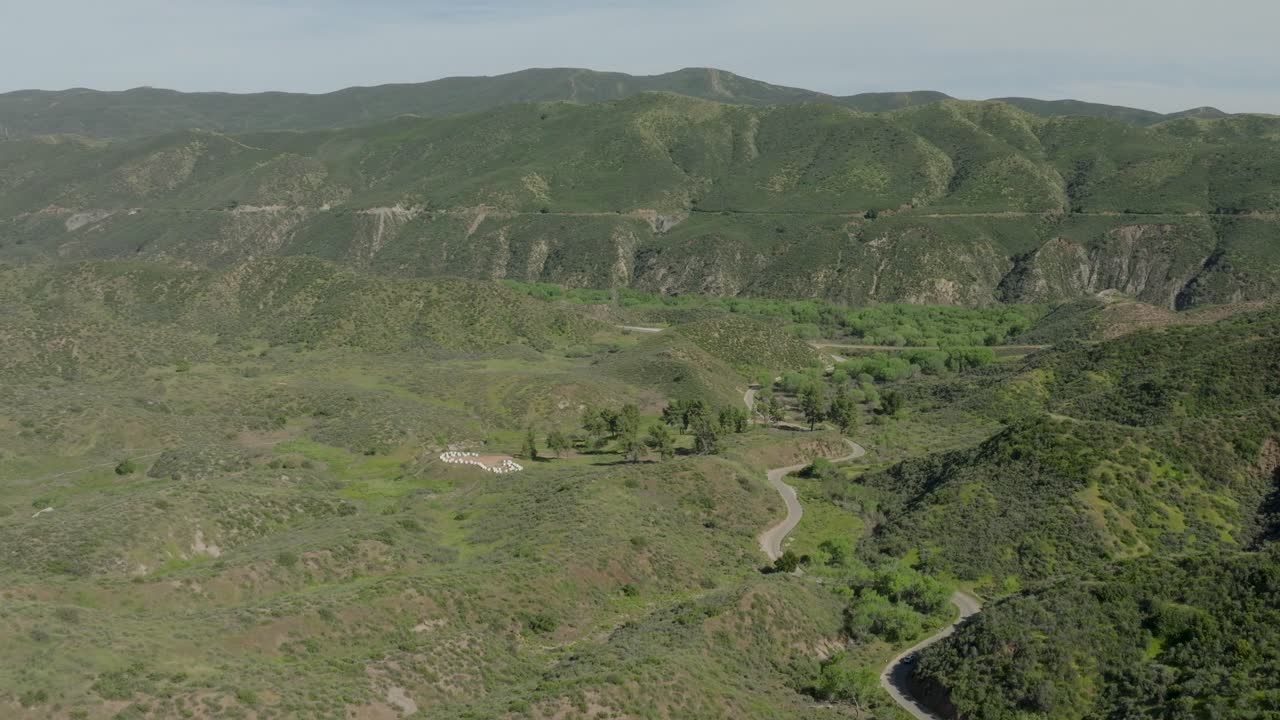 Aerial wide pull-out shot of a remote rural valley with dense green hills and a large beekeeping apiary on display next to a lonely winding country road.
