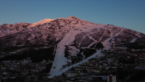 Luftaufnahme-Eines-Berges-Im-Sonnenuntergang-Mit-Skipiste-In-Der-Nähe-Einer-Stadt