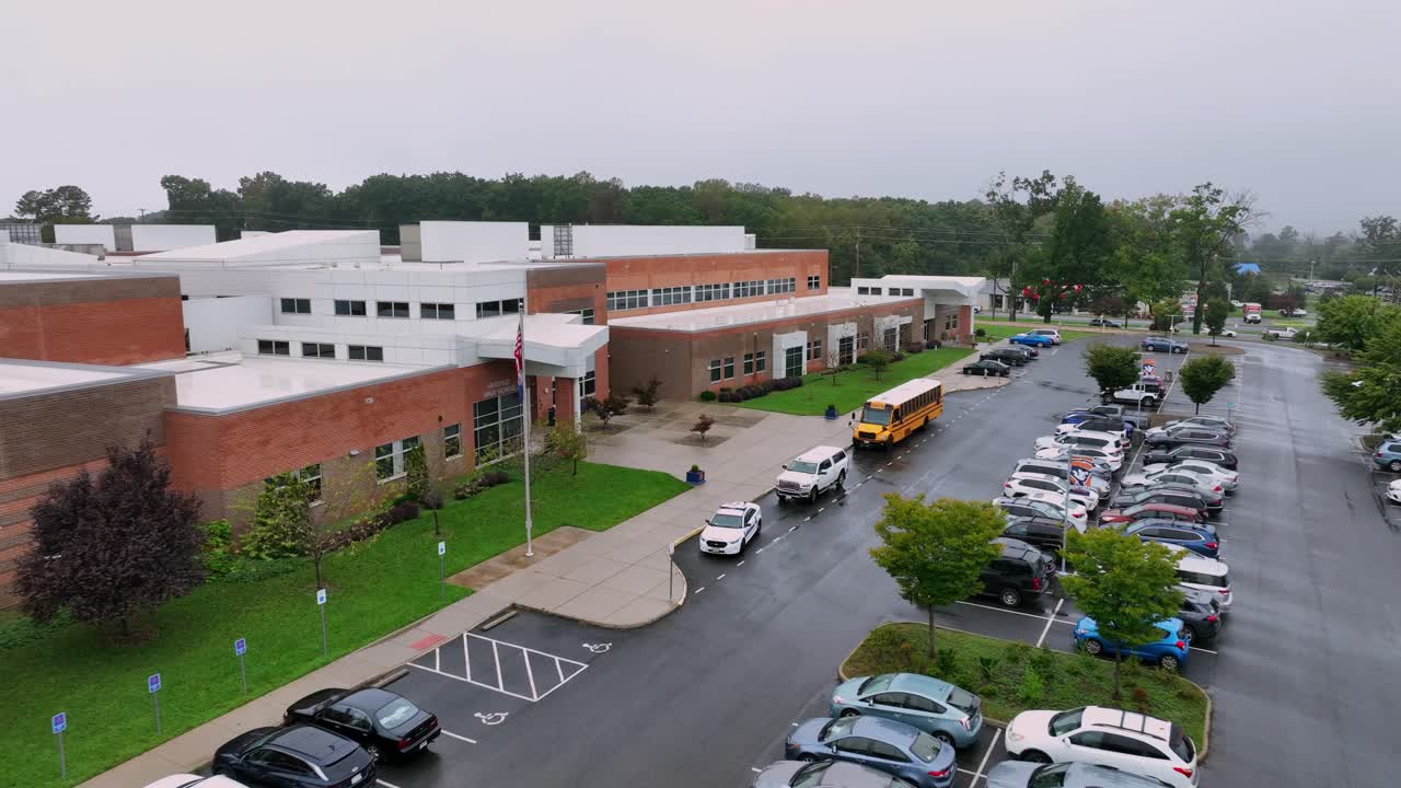 High school scene in American town during cloudy and rainy day. Parking yellow school bus in front of historic school of USA. Parking cars on parking lot.Driving cars on intersection road.Aerial view.